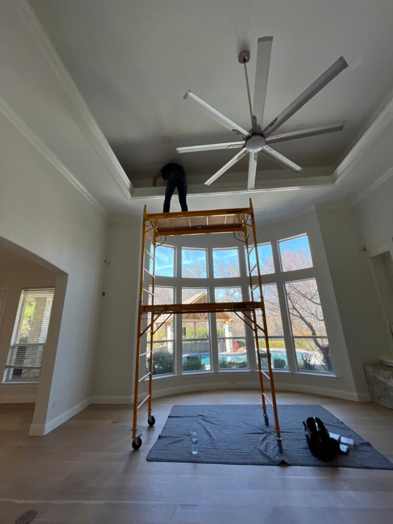 A worker standing on a scaffold painting or repairing a ceiling with a large fan in a well-lit living room