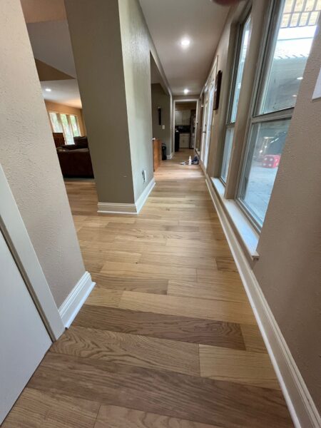 A view of a spacious hallway featuring new wooden flooring, with natural light streaming in from large windows.
